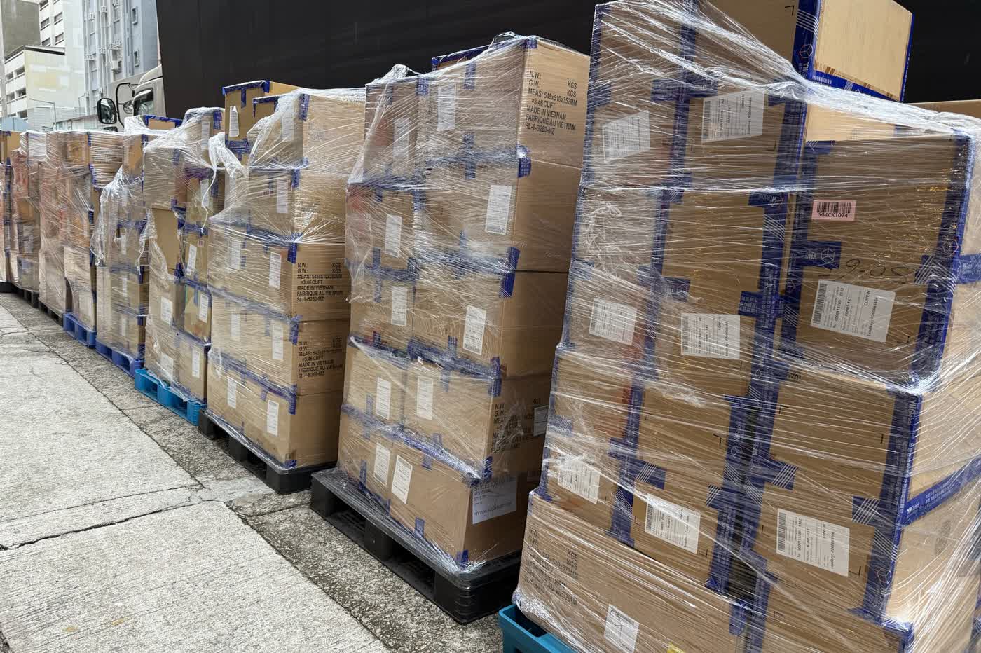 A row of shrink-wrapped pallets of sealed cartons lined up on a Hong Kong kerb beside a truck.
